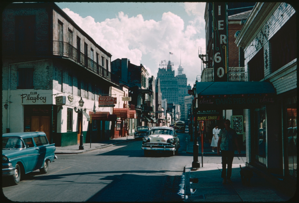 A vintage street scene with classic cars parked along the sidewalk. Buildings display signs for businesses, including a club and a pizza place. People walk under a shop awning, and a skyscraper is visible in the background under a cloudy sky.