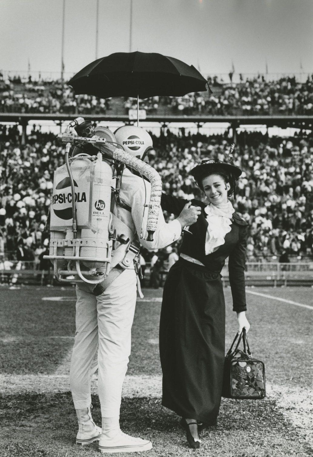 A person with a Pepsi-Cola jetpack and helmet holds an umbrella over a woman dressed in vintage clothing at a sports stadium. The crowd is seated in the background.