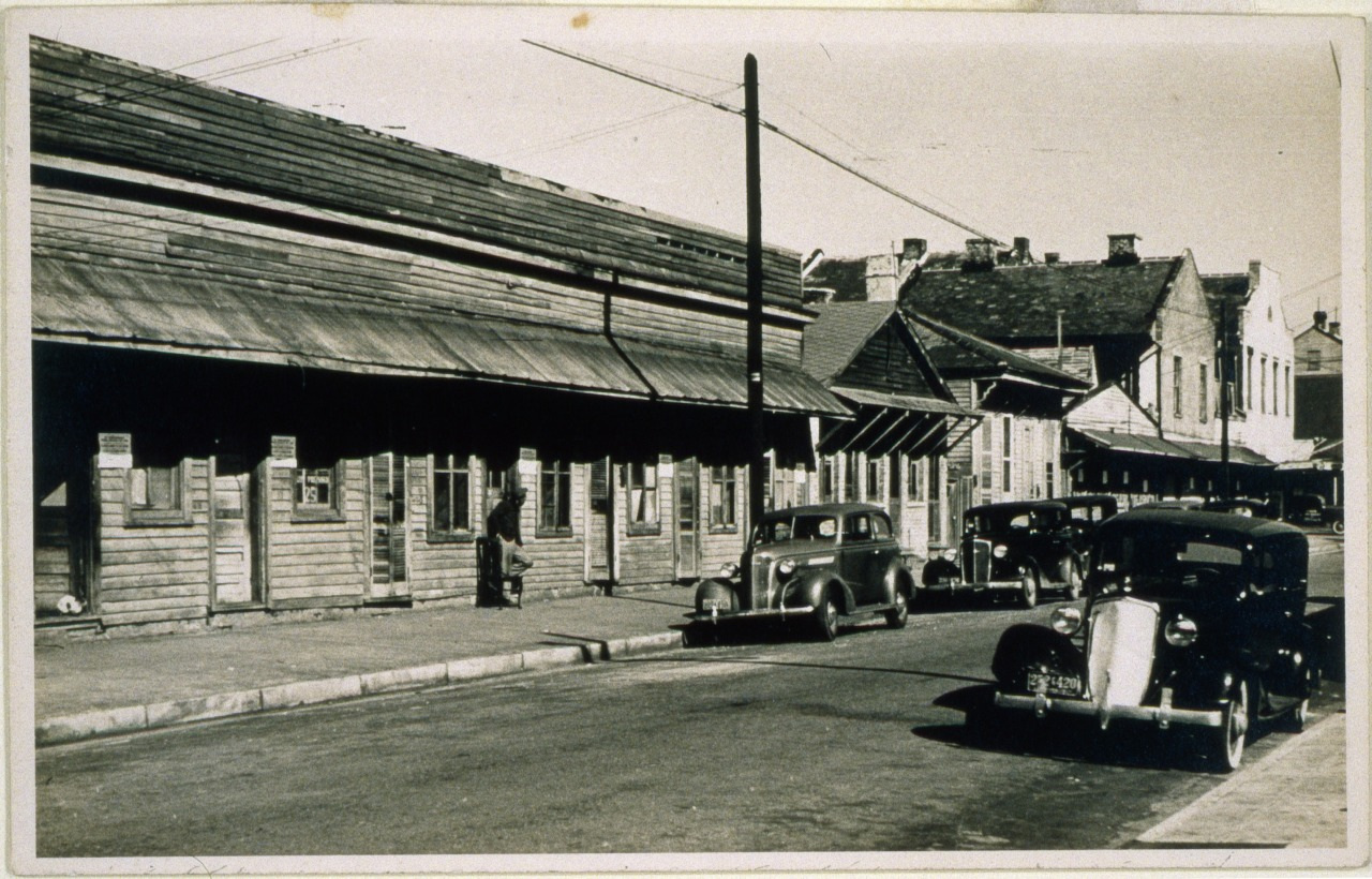A black-and-white photo shows a row of vintage cars parked along a street lined with old wooden buildings. A person stands near one of the buildings. The scene captures an early to mid-20th-century urban setting.