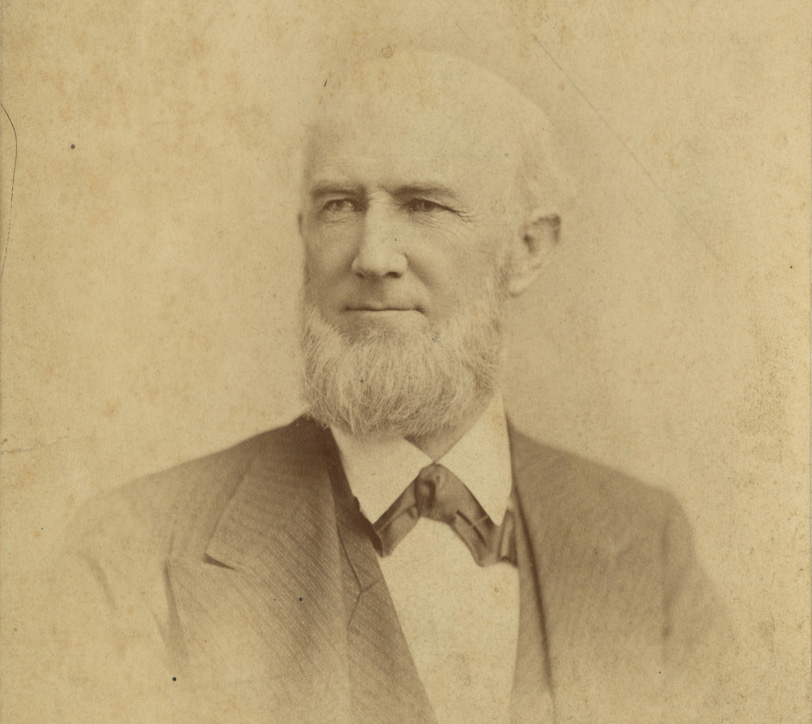 Sepia-toned portrait of an older man with a beard, wearing a suit and bow tie. The image has an antique feel with slight discoloration, suggesting age.