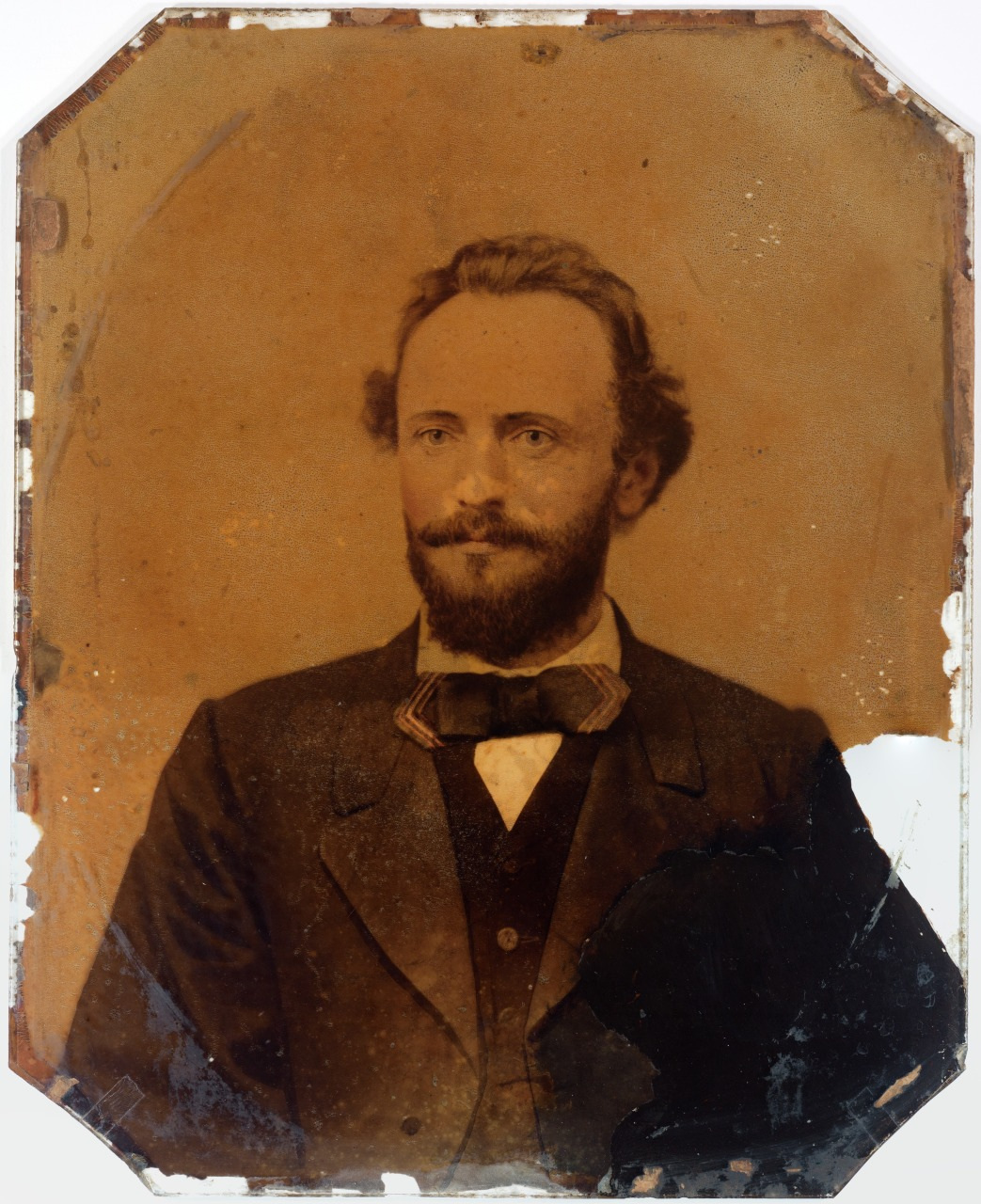 A sepia-toned vintage portrait of a man with a beard and mustache, wearing a dark suit with a bow tie. The image is worn and has visible scratches and edges softened by age.