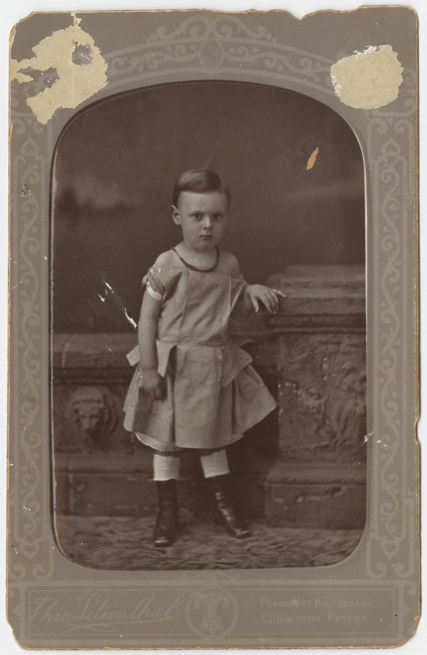 A sepia-toned vintage photograph of a young child with short hair, wearing a dress and boots, standing with one hand resting on an ornate stone pedestal. The photo is framed with decorative detailing at the edges.