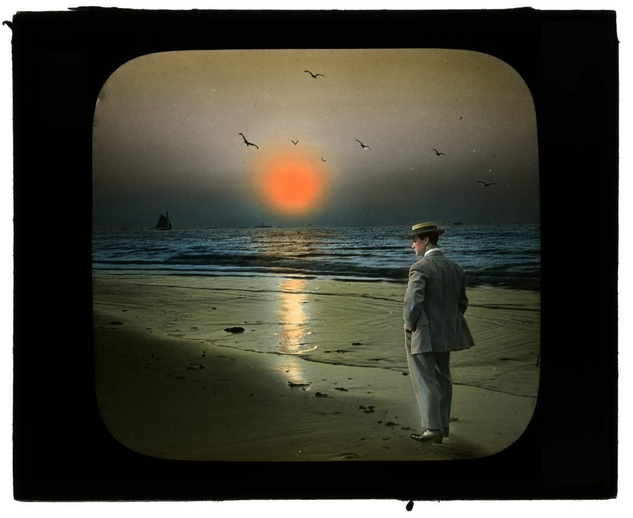 A man in a suit and hat stands on a beach, gazing at a serene sunset over the ocean. Birds fly in the sky, and a sailboat is visible on the horizon. The sky has hues of orange and blue reflected on the water.
