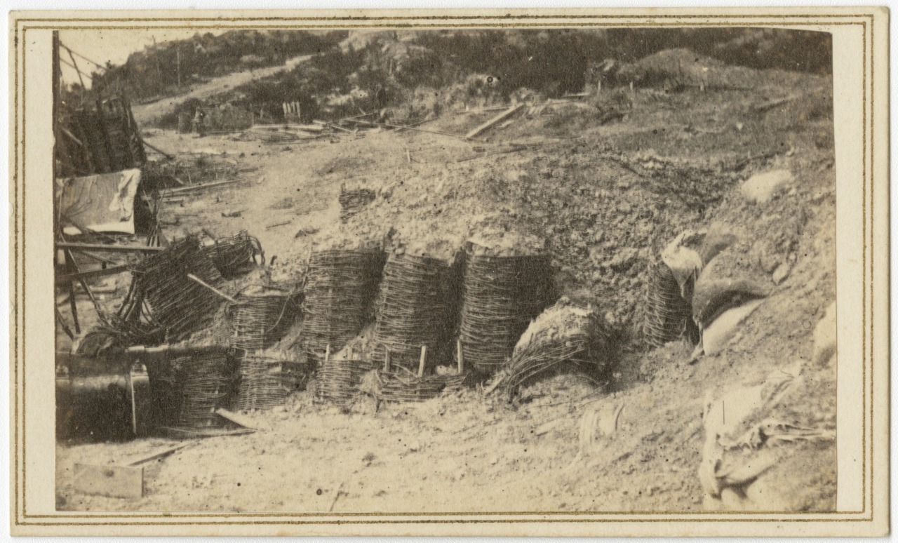 A sepia-toned photograph shows a rugged terrain with several large bundles of twigs or branches stacked against an embankment. The landscape is sparse, and there are wooden structures in the background.