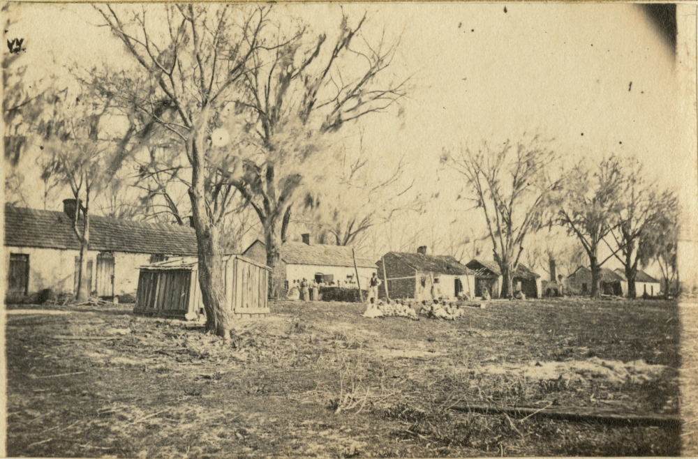 A sepia-toned photograph shows several small, rustic buildings lined up on a barren landscape with leafless trees. People are gathered in front of one building, and wooden structures are visible nearby. It has an old, historical appearance.