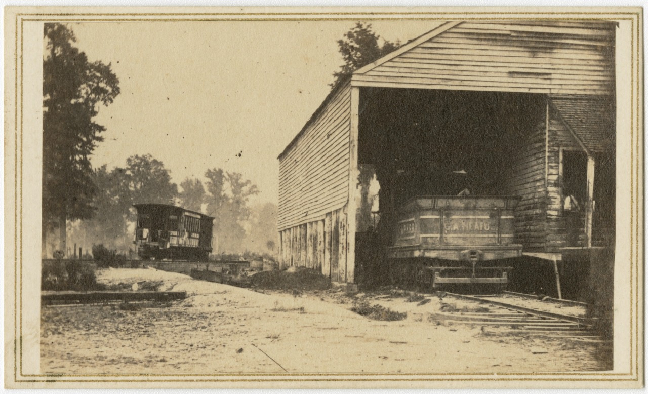 A vintage photograph shows a train with a caboose entering a wooden shed on a dirt road, surrounded by trees. Another part of the train is visible on the left side. The image is sepia-toned, capturing a rural scene.
