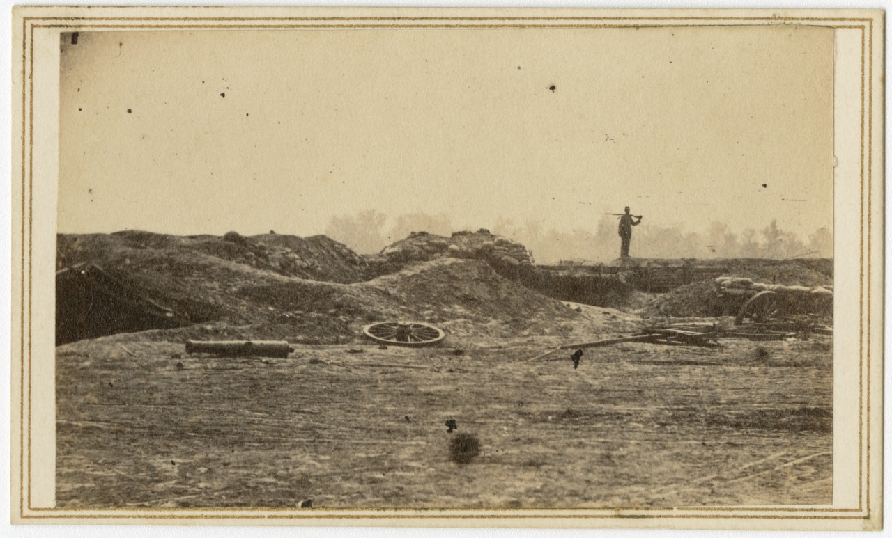 A sepia-toned photograph of a barren battlefield with artillery debris scattered on the ground. A lone figure stands on the horizon against a backdrop of trees and a hazy sky, surveying the terrain through a looking glass.