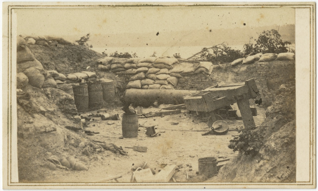 A black and white photograph depicts a military fortification, featuring a cannon surrounded by sandbags. Various barrels and wooden debris are scattered around the trench. In the background, trees and a body of water are visible.
