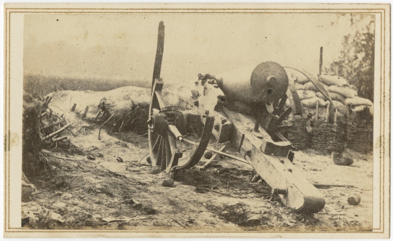 A vintage photo of an old-fashioned cannon positioned on rough terrain, surrounded by sandbag fortifications. The cannon is oriented towards the left, with scattered debris and trees visible in the background.