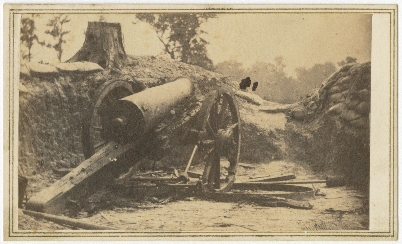 A vintage photograph of an old, worn-out cannon on large wooden wheels, positioned on rough terrain. The cannon is surrounded by logs and sandbags, suggesting a historical battlefield.