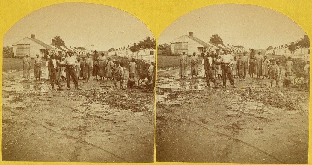 A vintage stereoscopic photo shows a group of people, including several children, gathered in an outdoor area with puddles. Buildings and trees are visible in the background. Some individuals stand while others sit or crouch on the ground.