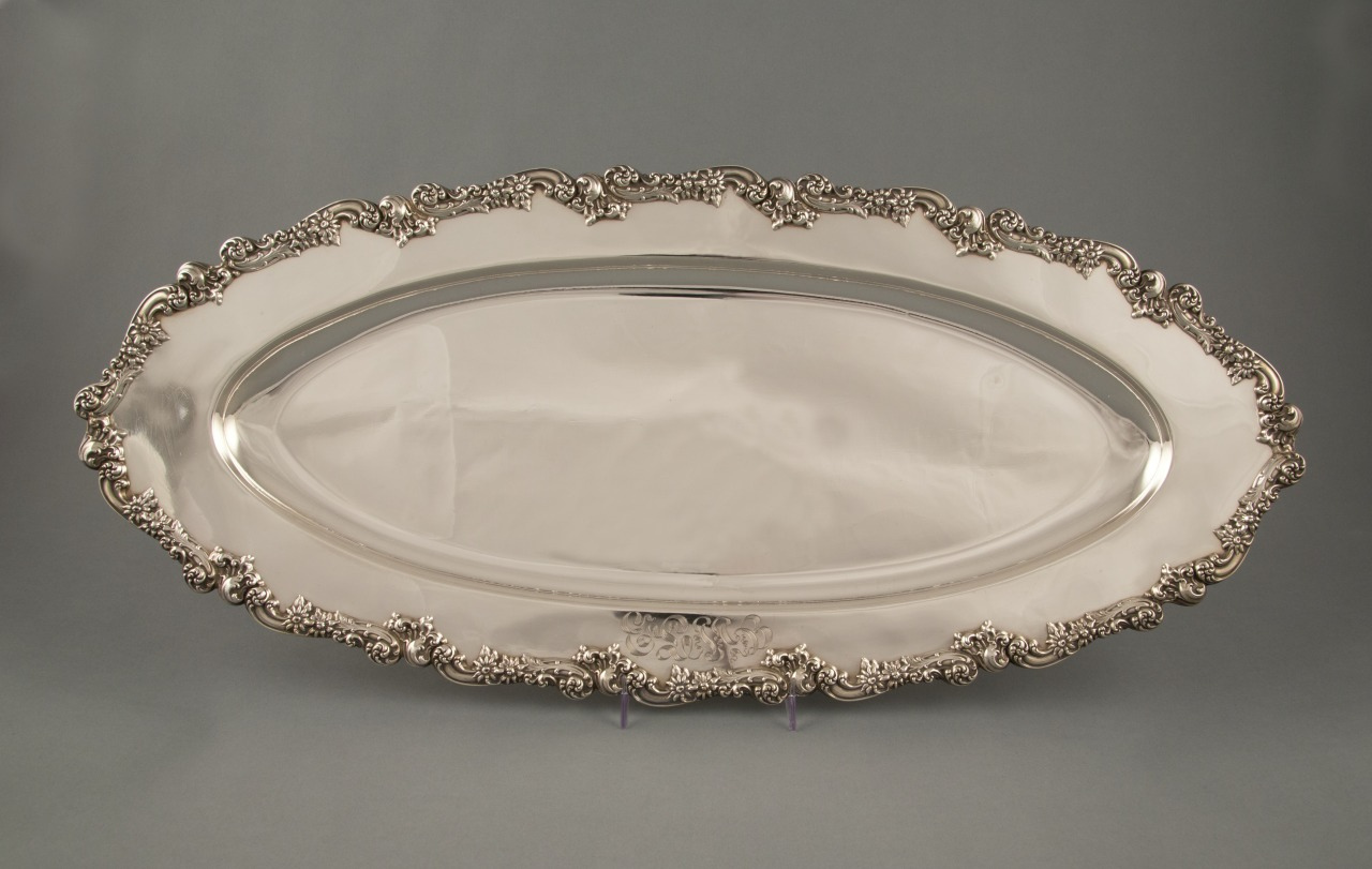 An ornate, oval-shaped silver platter with intricate floral patterns along the edges against a plain gray background.