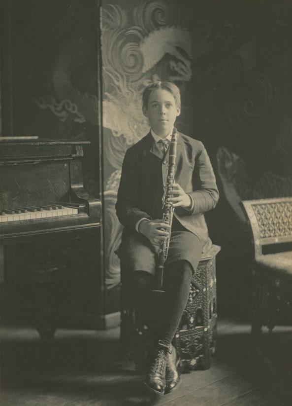 A young person in formal attire sits on a decorative stool holding a clarinet. They are beside a piano with an ornate backdrop featuring swirling patterns, possibly dragons, creating an elegant and artistic atmosphere.