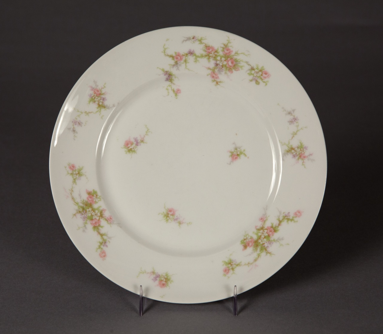A delicate white ceramic plate with a floral design featuring small pink flowers and green leaves. The plate is displayed on a clear stand against a dark background.