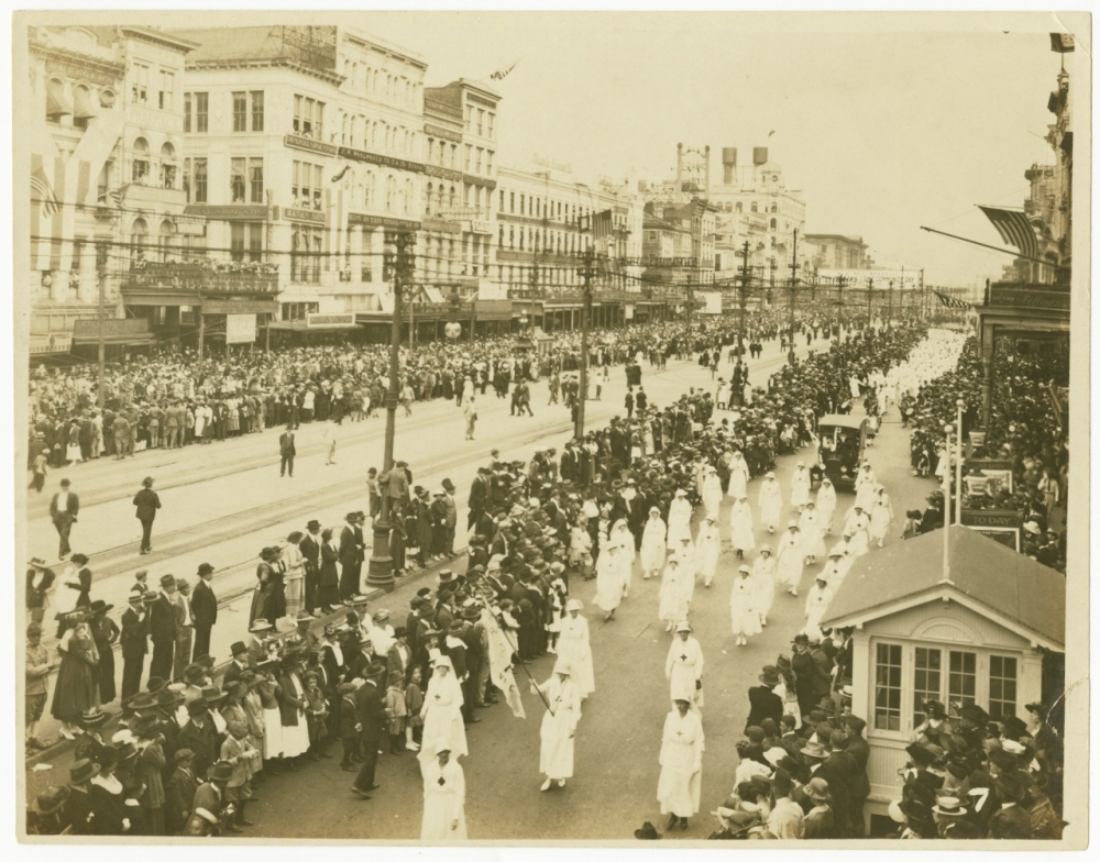 A historic photograph showing a large parade with participants in white hooded robes walking down a wide street lined with spectators. The buildings in the background are vintage, and the crowd watches intently from both sides.