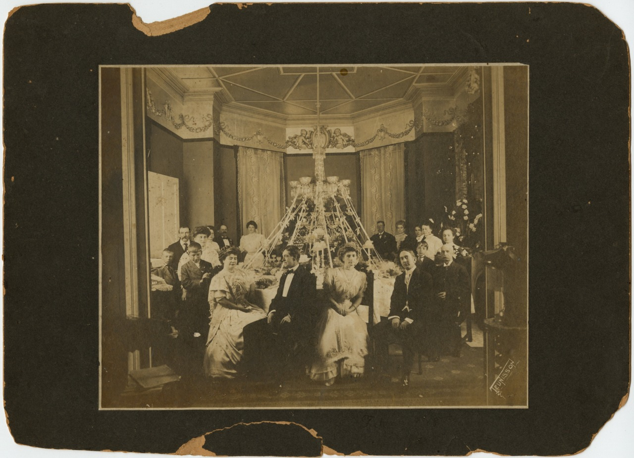A vintage sepia-toned photograph depicting a group of people seated around a grand dining table, adorned with an elaborate centerpiece. The setting appears formal, with men in suits and women in elegant dresses. The room is richly decorated.