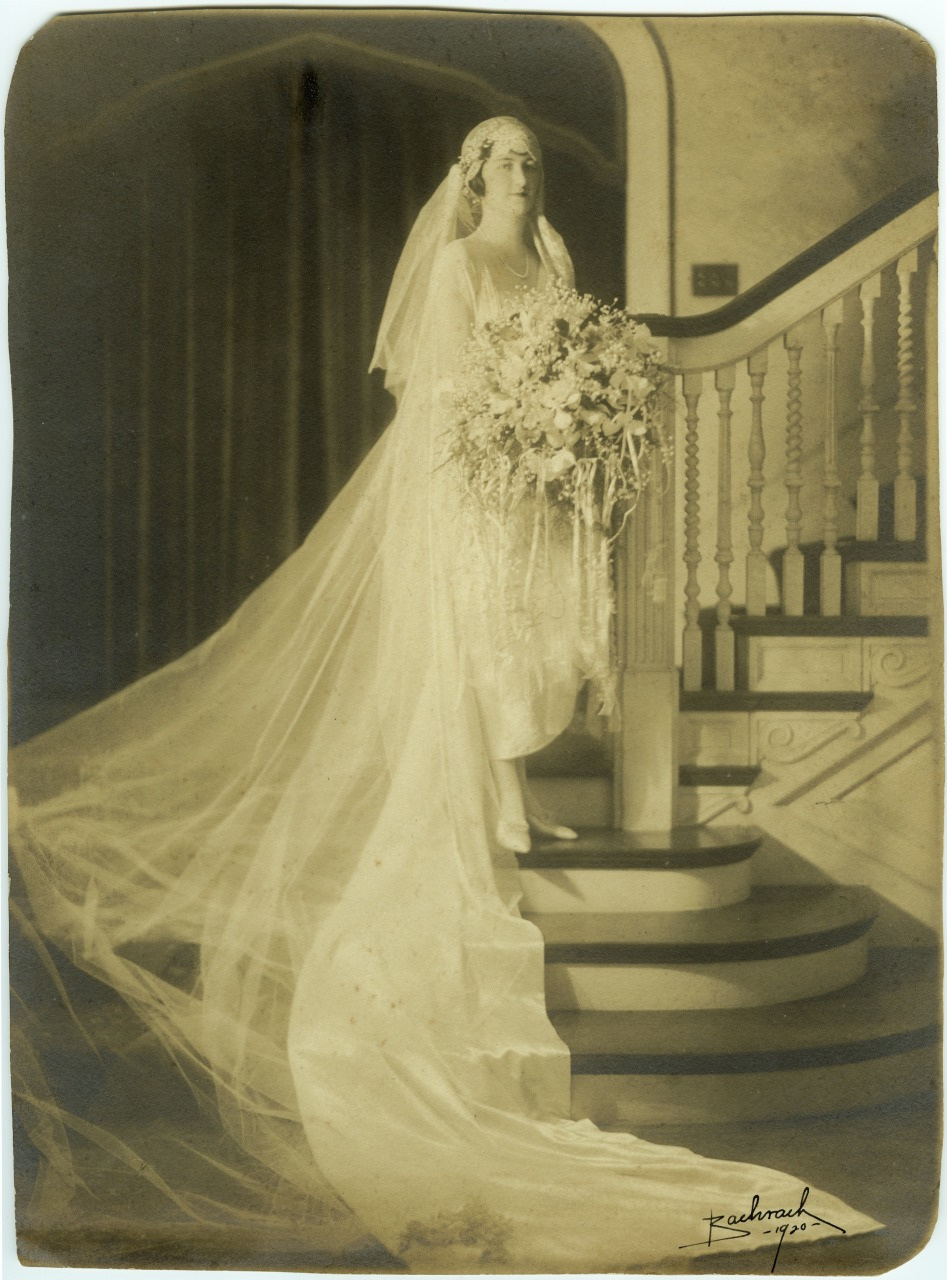A vintage sepia-toned photograph of a bride in a long, flowing wedding dress and veil, holding a large bouquet. She stands on a staircase with ornate wooden railings, exuding an elegant and timeless feel.