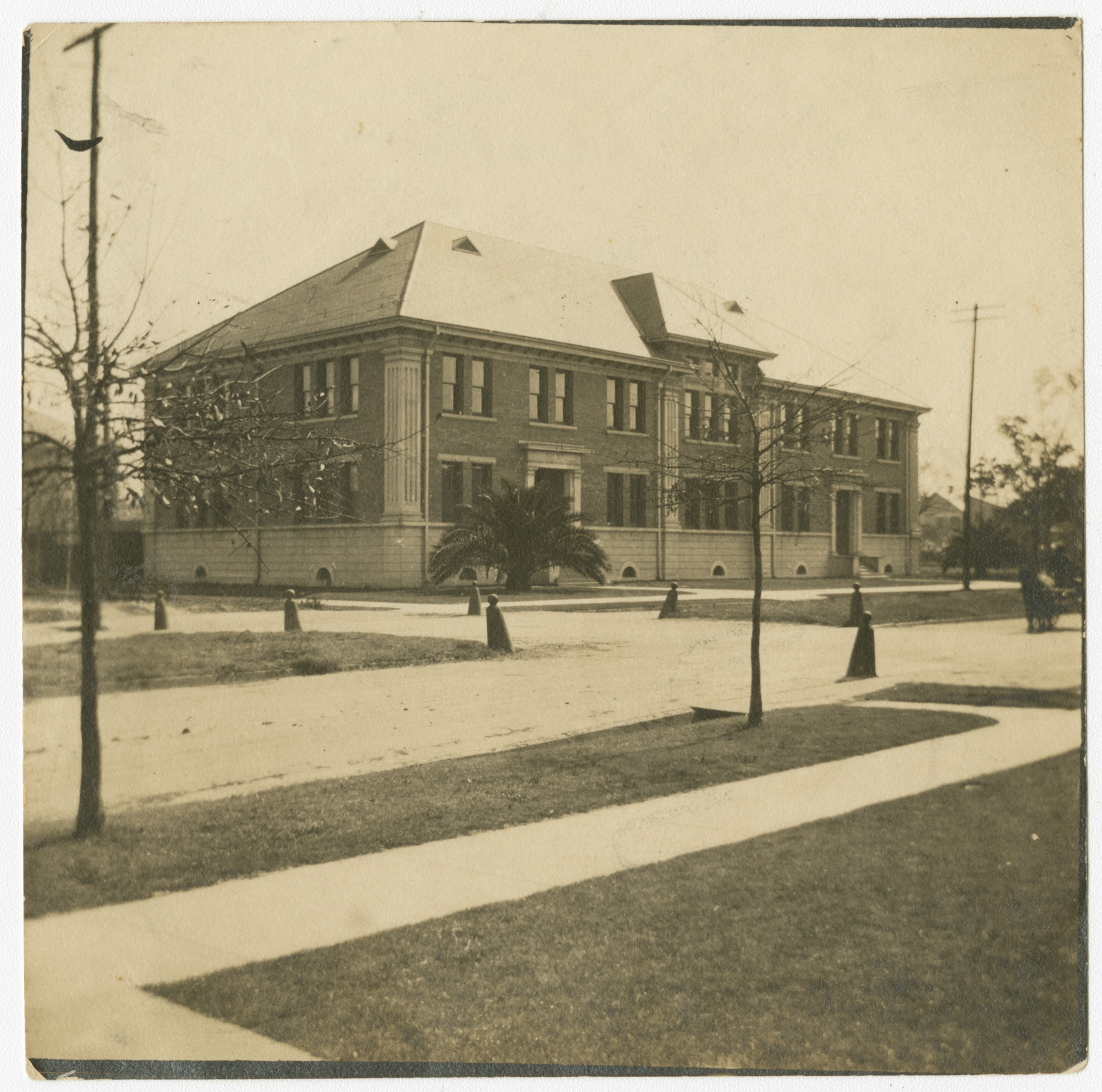 A sepia-toned photo of a two-story brick building with multiple windows, set on a corner with paved sidewalks and surrounded by small trees and grass. Vintage aesthetics suggest an early 20th-century architectural style.