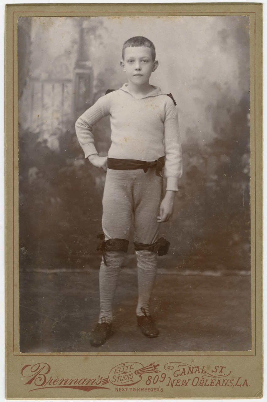 A young boy in vintage attire stands confidently with one hand on his hip. He wears a sweater, knee-length shorts, and high socks, with bows on his knees. The photo is from Brennans Elite Studio in New Orleans, as noted in the bottom text.