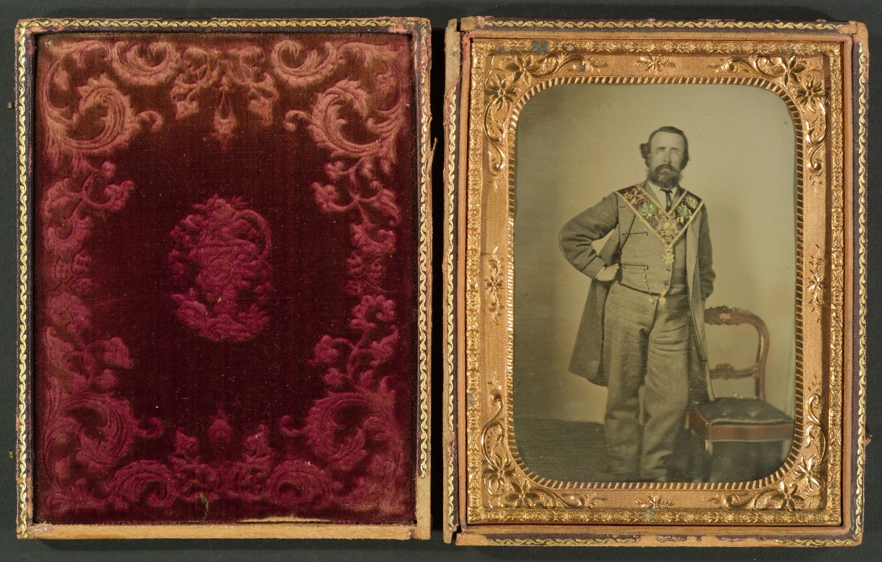 A sepia-toned portrait of a man in 19th-century formal attire, standing beside a chair. The image is framed in an ornate gold case with a deep red, velvet-like interior design on the left side.
