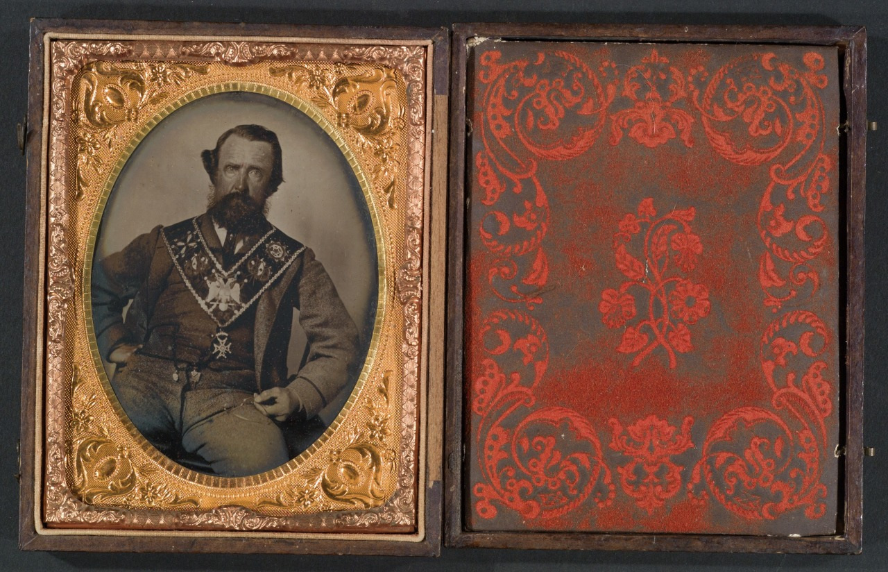 A vintage photograph of a bearded man in ornate clothing, seated, enclosed in an intricate gold frame. The case is open, revealing decorative red patterns on the opposite side.