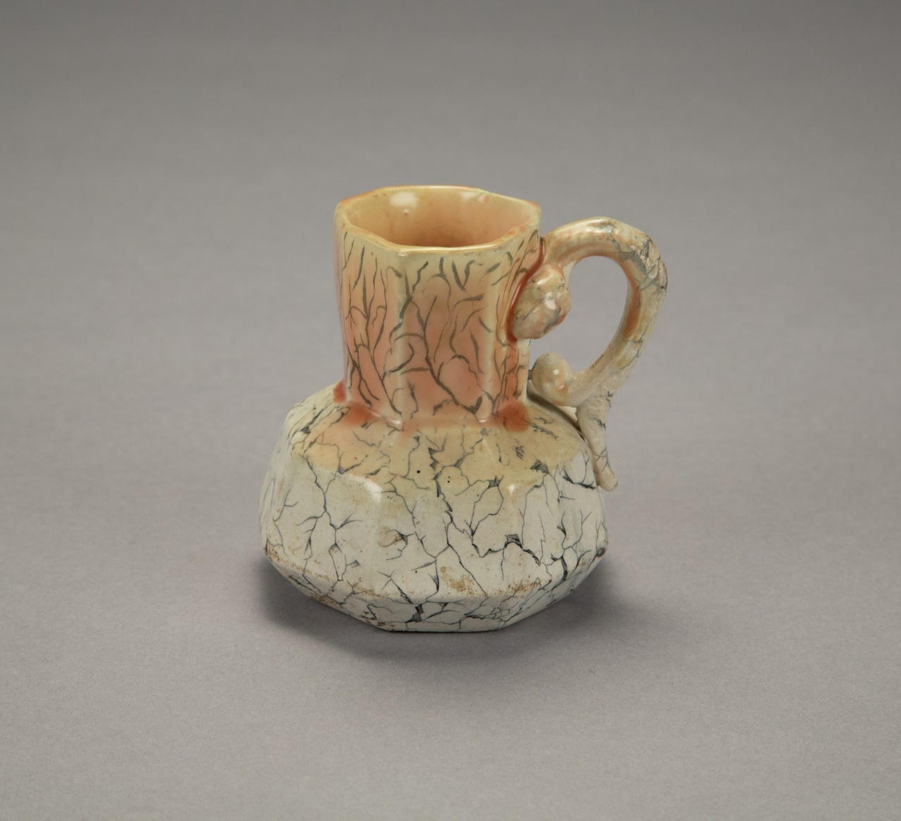 A ceramic mug with a textured, crackled finish sits on a gray surface. The mug features a thick handle and a bulbous base, with colors ranging from peach to grayish tones, showcasing a unique and artistic design.