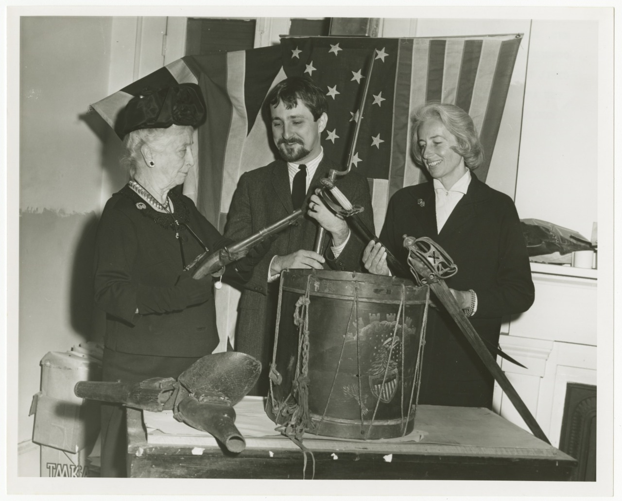 Three people standing behind a table displaying historical artifacts, including a musket and a drum. They are dressed in formal attire. An American flag is draped in the background.