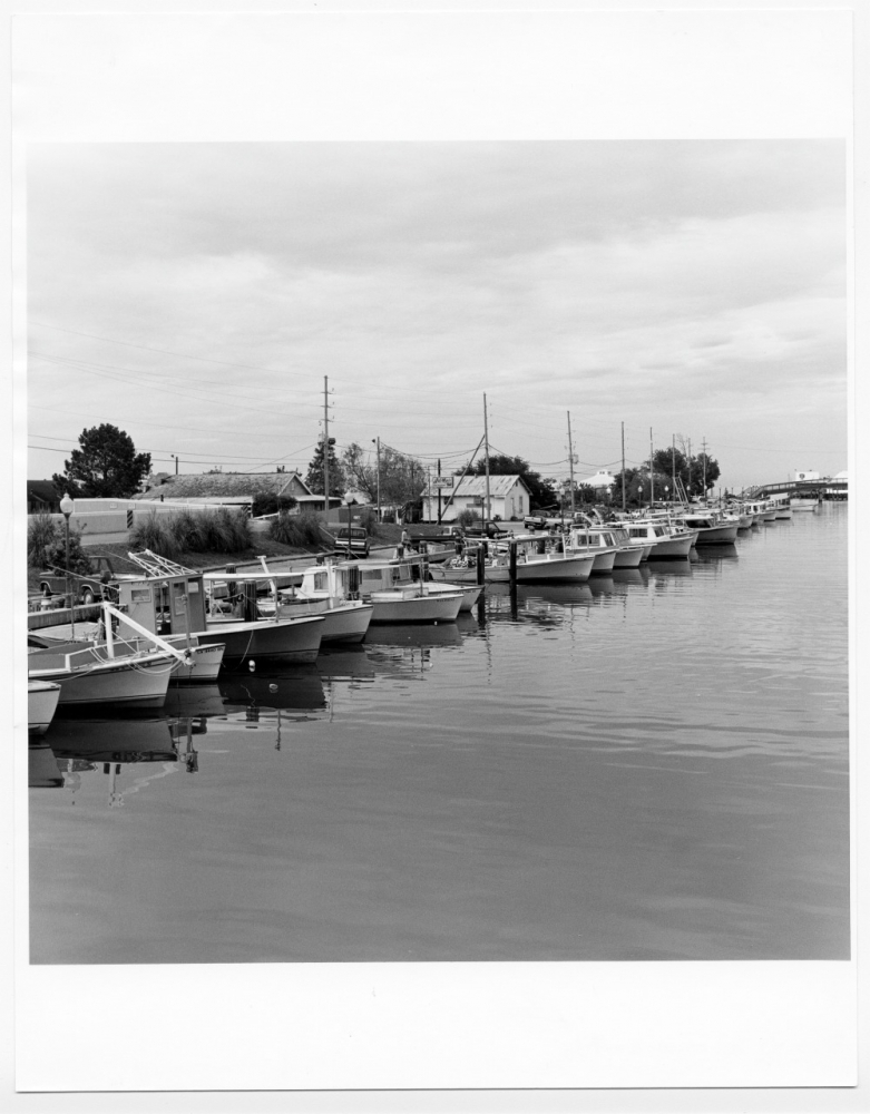 Black and white photo of a marina with several docked boats along a calm waterway. Buildings and trees are visible in the background under a cloudy sky.