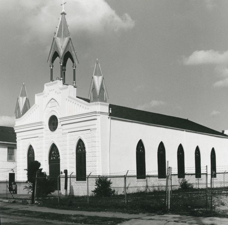 A black and white photo of a Gothic Revival style church with pointed arches and a central tower. The building is surrounded by a chain-link fence, with a few trees and bushes nearby. The sky is partly cloudy.