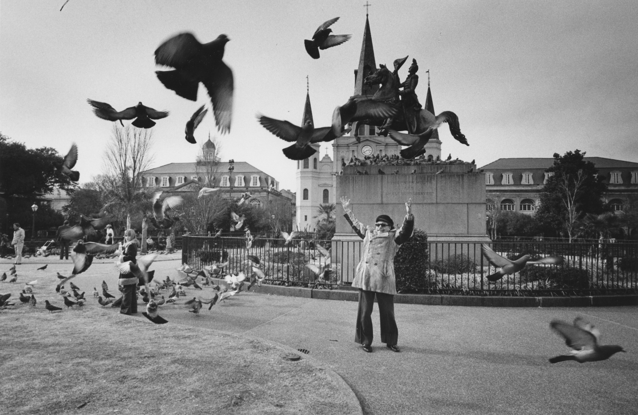 A person stands in a park with arms raised, surrounded by flying pigeons and more on the ground. In the background, a historical building with tall spires and a clock is visible, along with other people and trees. The scene is lively and dynamic.