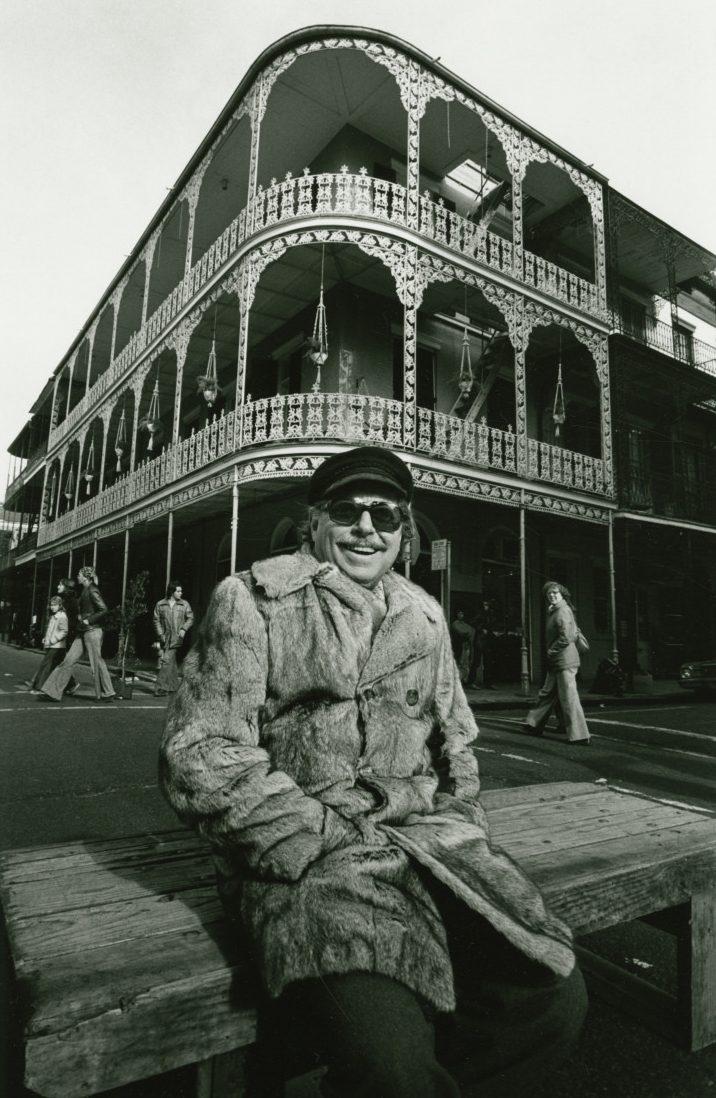 A person wearing a fur coat and sunglasses sits on a wooden bench in front of an ornate, multi-story building with wrought-iron balconies. People walk on the street nearby.