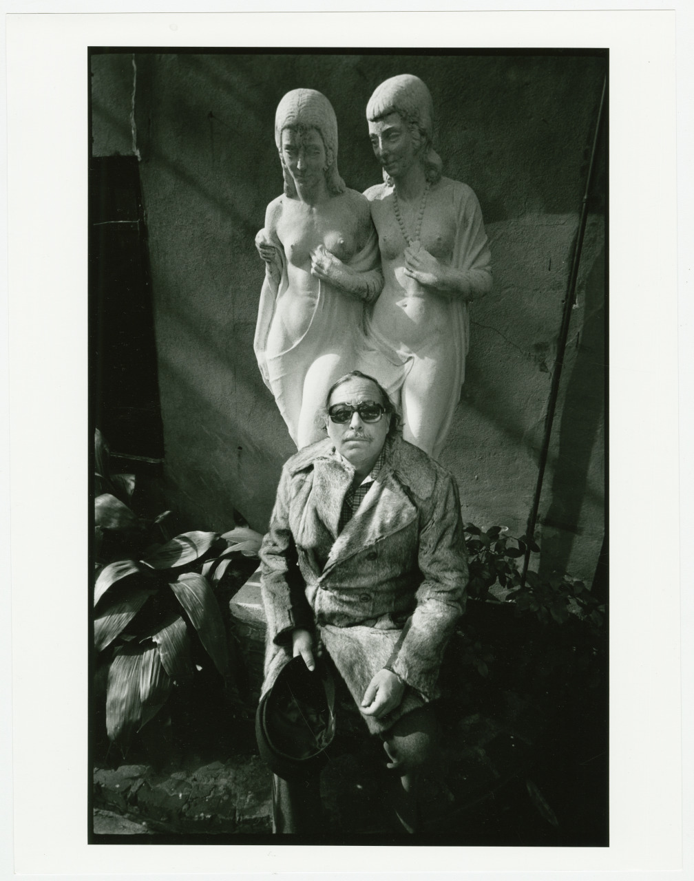 A person wearing sunglasses and a coat sits on a bench, holding a hat in their lap. Behind them is a sculpture of two classical figures standing close together. The scene is outdoors with shadows cast on the wall.