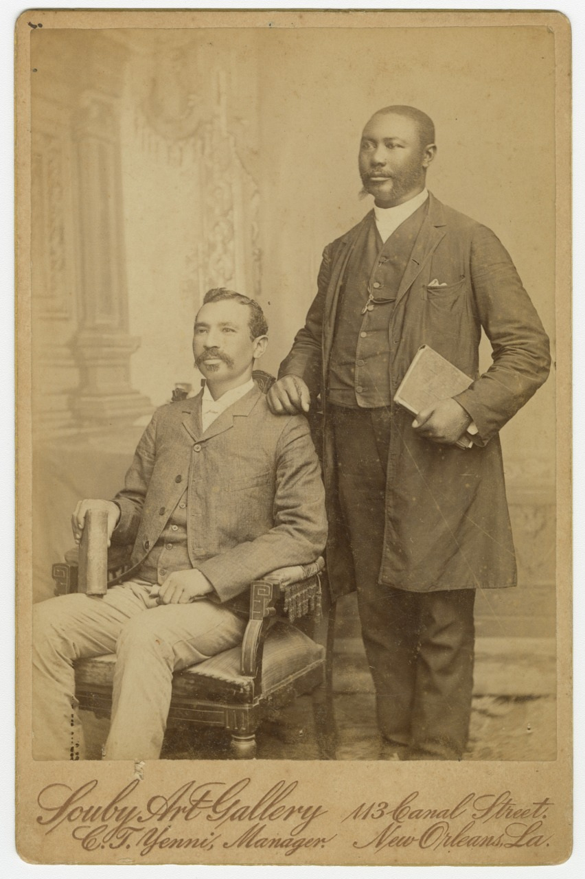 Two men pose in a vintage photograph. One is seated, holding a scroll, wearing a suit with a bow tie. The other stands, resting a hand on the chair, holding a book, dressed in a suit with a boutonnière. The studio name appears at the bottom.