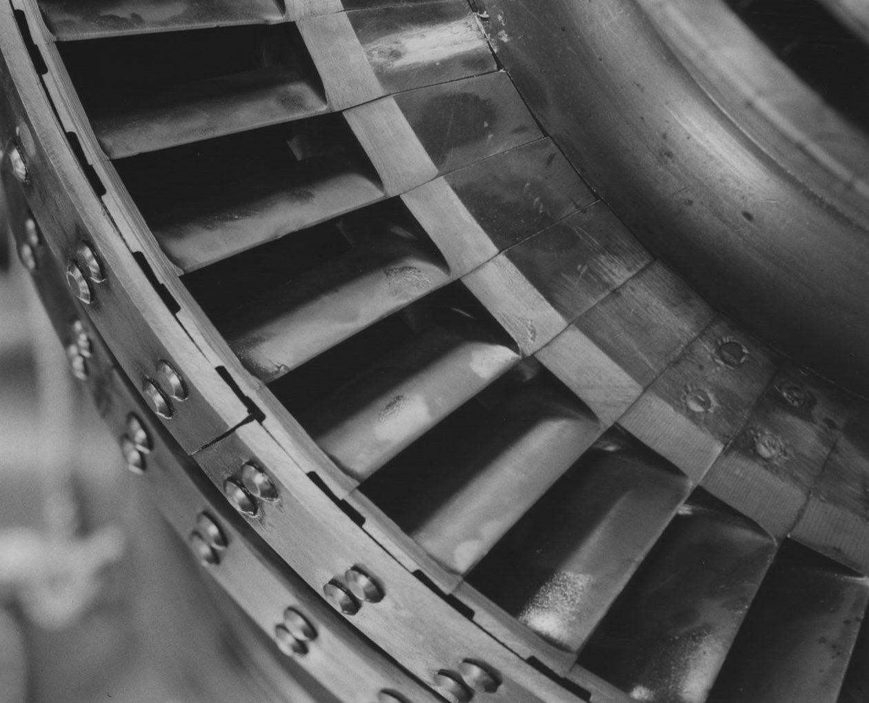Close-up of a turbines metal blades arranged in a circular pattern. The surface appears smooth and shiny, with rivets visible along the edge. The blades have a slightly angled design, suggesting motion or airflow.