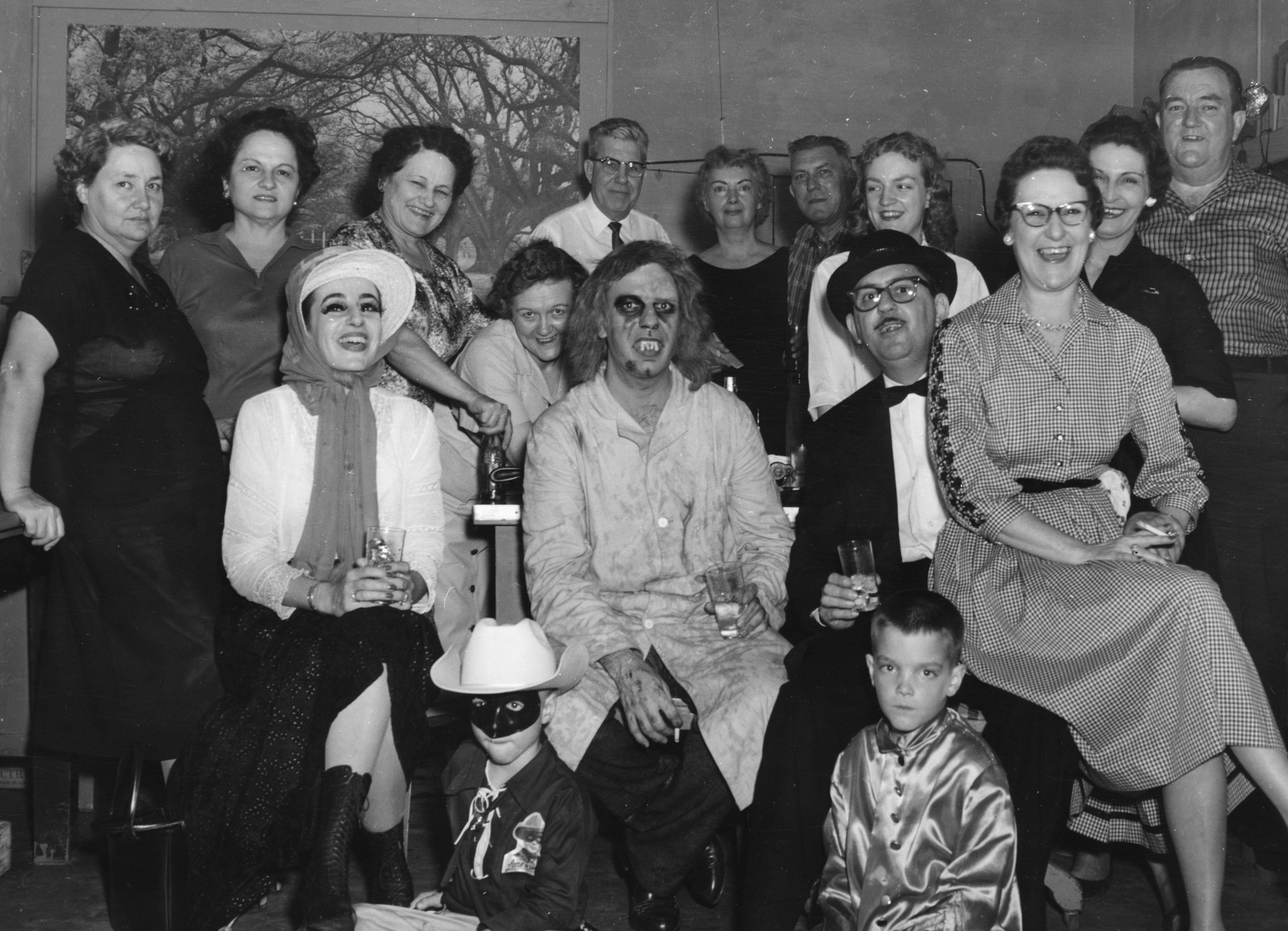 A group of people in a vintage setting, some in costume. A person dressed as a cowboy kneels in front. Others wear outfits like robes and hats. They are gathered closely, smiling, with drinks in hand, in what seems to be a casual gathering.