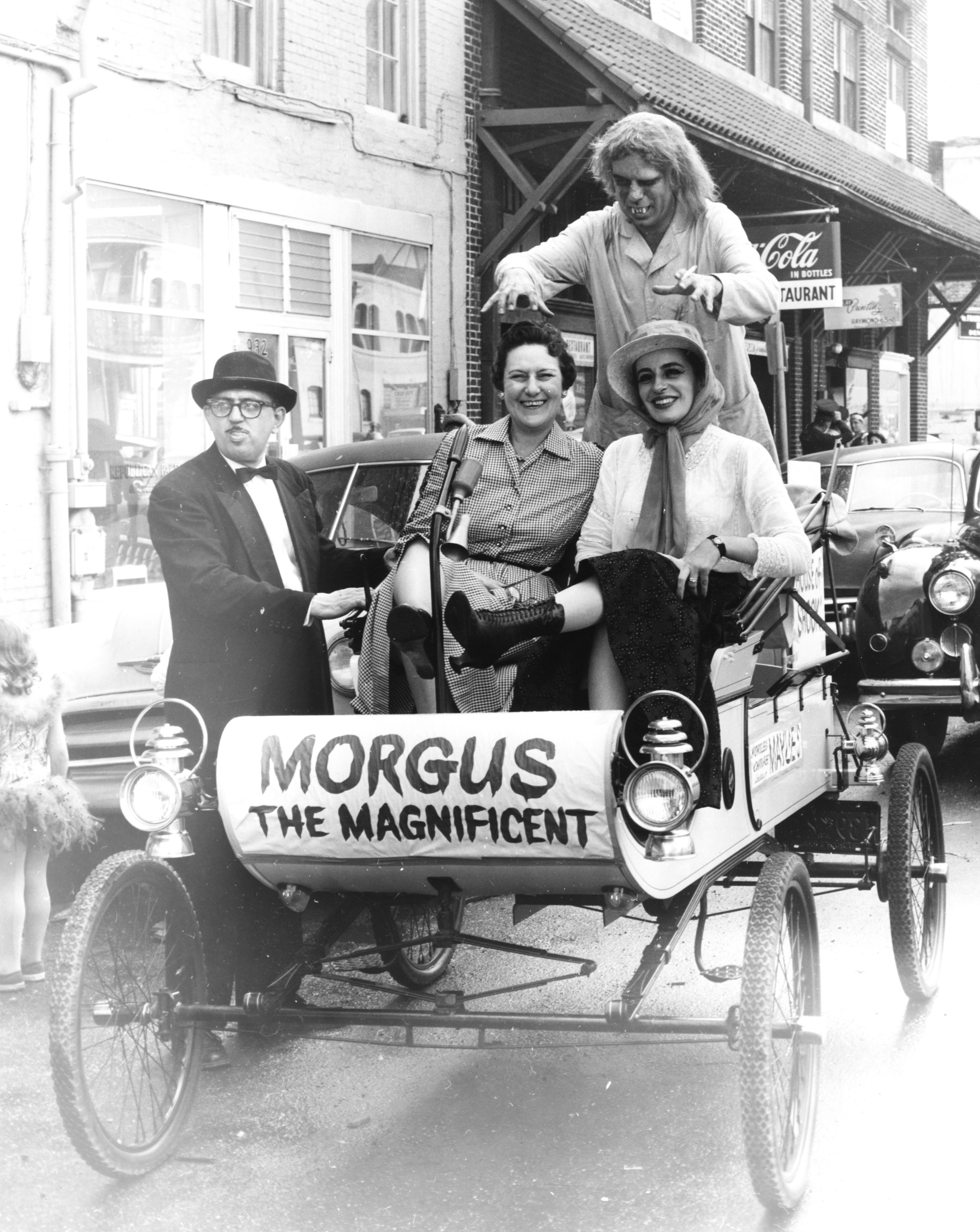 A black-and-white photo depicts two women sitting in an antique car labeled Morgus the Magnificent, with a man driving and another man standing, dressed as a monster, reaching over them. They are on a street with vintage cars and a Coca-Cola sign in the background.