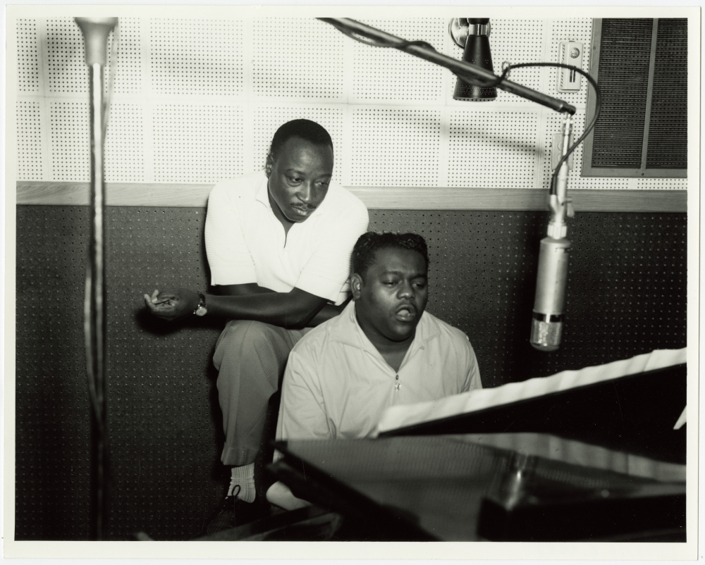 Two men are at a recording studio. One man sits at a piano, singing, while the other kneels on a chair beside him, listening attentively. A large microphone is suspended above them. The wall is covered with acoustic panels.