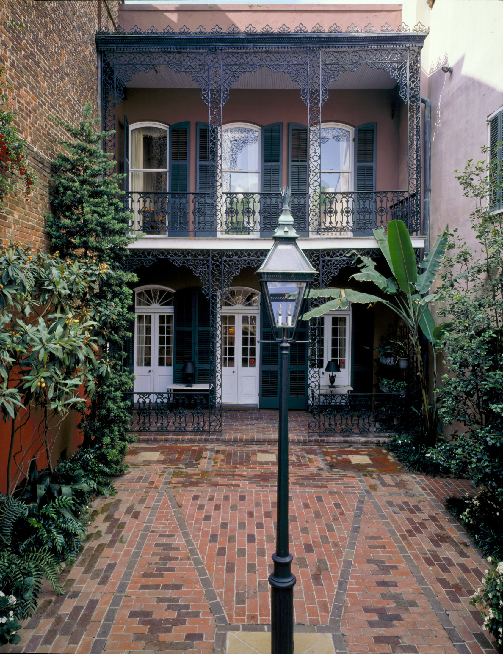A charming courtyard with brick flooring features a traditional black lamppost in the center. Surrounding the area are lush green plants, and behind is a two-story building with ornate wrought-iron balconies and tall windows.