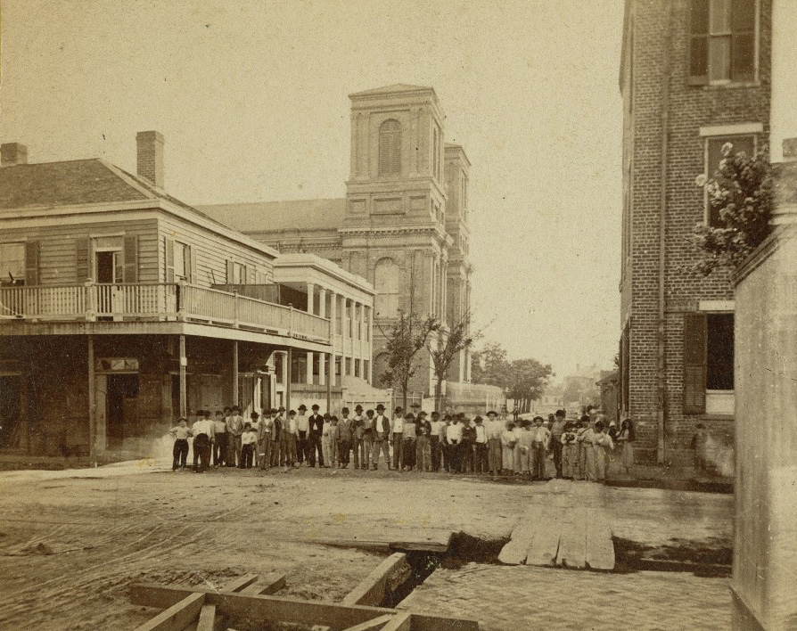 A vintage photograph depicts a group of people, including men, women, and children, standing on a dirt road surrounded by buildings. The scene includes wooden sidewalks and a church tower in the background.