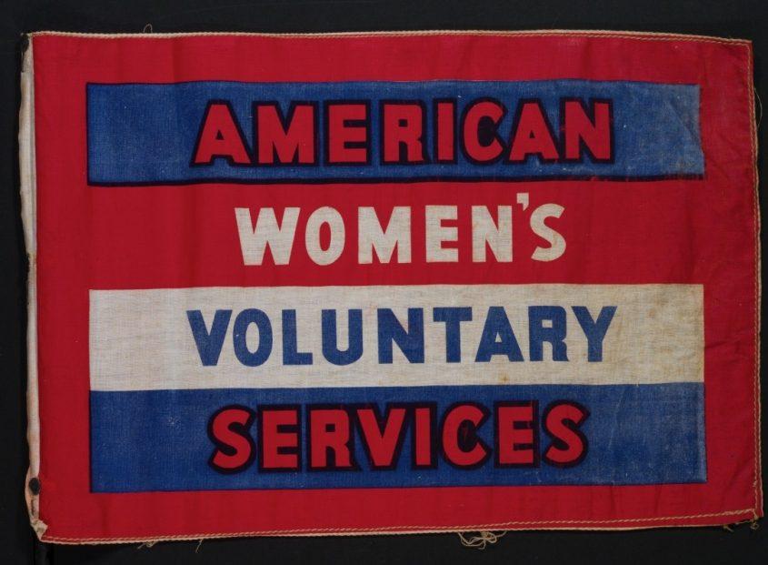 A rectangular flag with a red background and three horizontal sections. The text reads AMERICAN WOMENS VOLUNTARY SERVICES in bold letters, with each word on a separate colored stripe of red, white, and blue.
