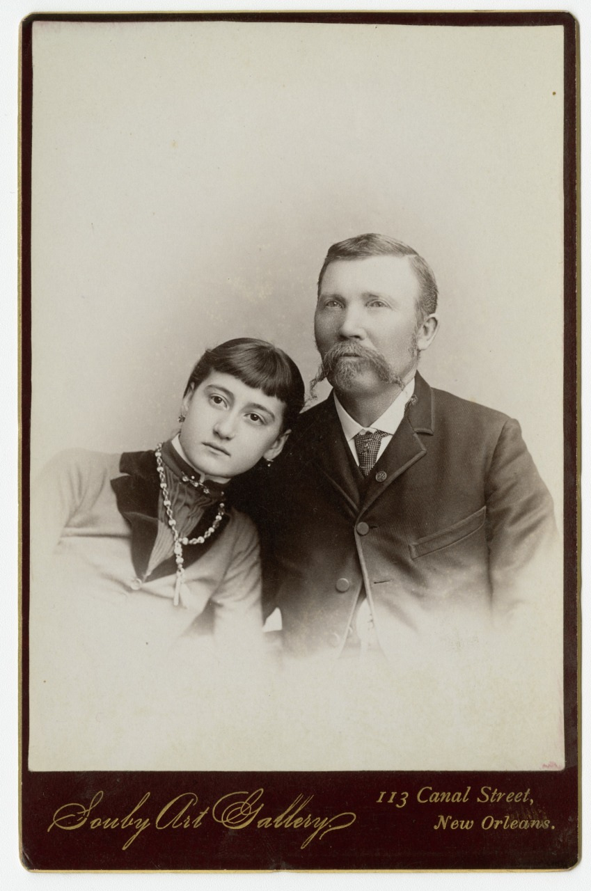 A vintage portrait of a woman leaning her head on a mans shoulder. The man has a mustache and wears a suit, while the woman wears a dress with a high collar and a necklace. The photo is from Soudy Art Gallery, New Orleans.