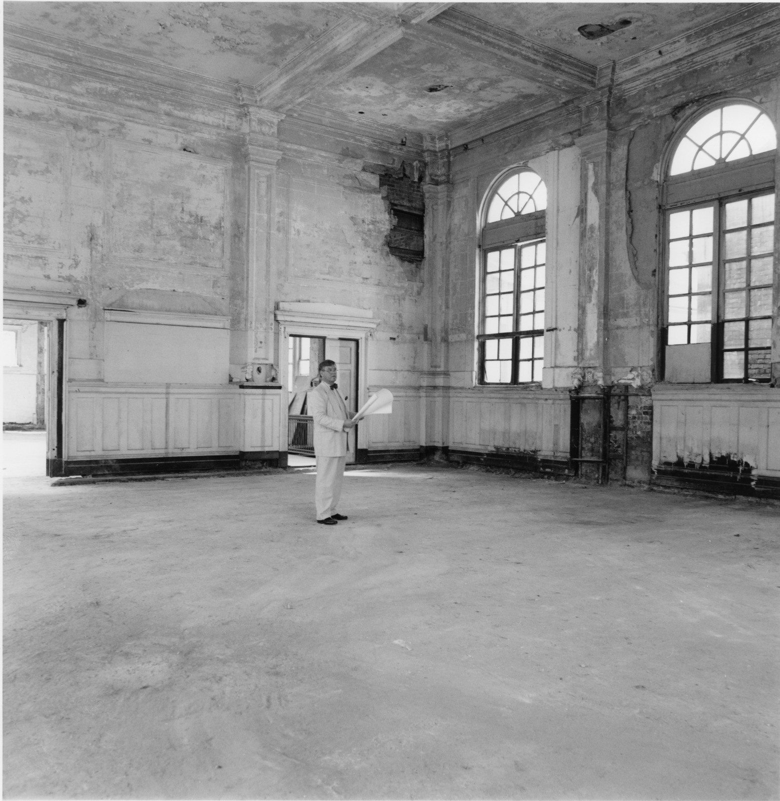 A person in a suit stands alone in a large, empty, and dilapidated room with high ceilings, large arched windows, and worn walls. The person appears to be inspecting or surveying the area while holding a rolled-up document.