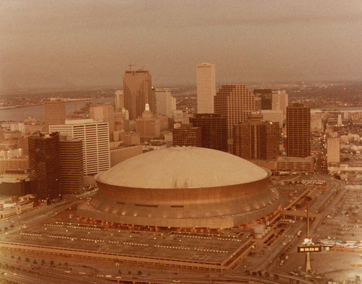 Aerial view of a dome-shaped stadium in an urban setting, with tall buildings in the background. The scene appears to be at dusk, with an orange tint over the landscape. Various city lights and roads are visible surrounding the stadium.