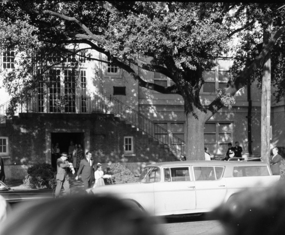 A black and white photo shows several people walking on a street in front of a large building. A long car is parked on the road, and a few individuals are near it. The scene is surrounded by large trees, casting shadows on the ground.