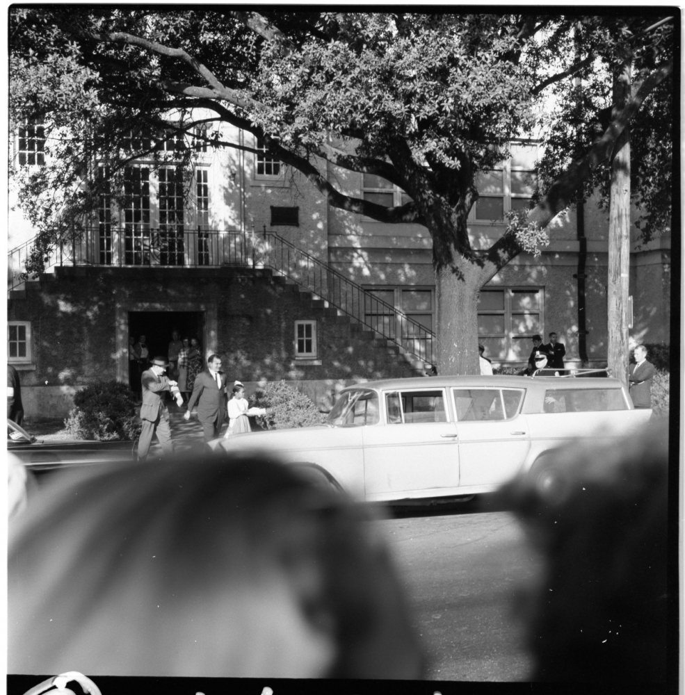 A black and white photo of a street scene with a vintage car parked in front of a large building. People are walking on the sidewalk, and a large tree provides shade. Blurred heads in the foreground slightly obstruct the view.