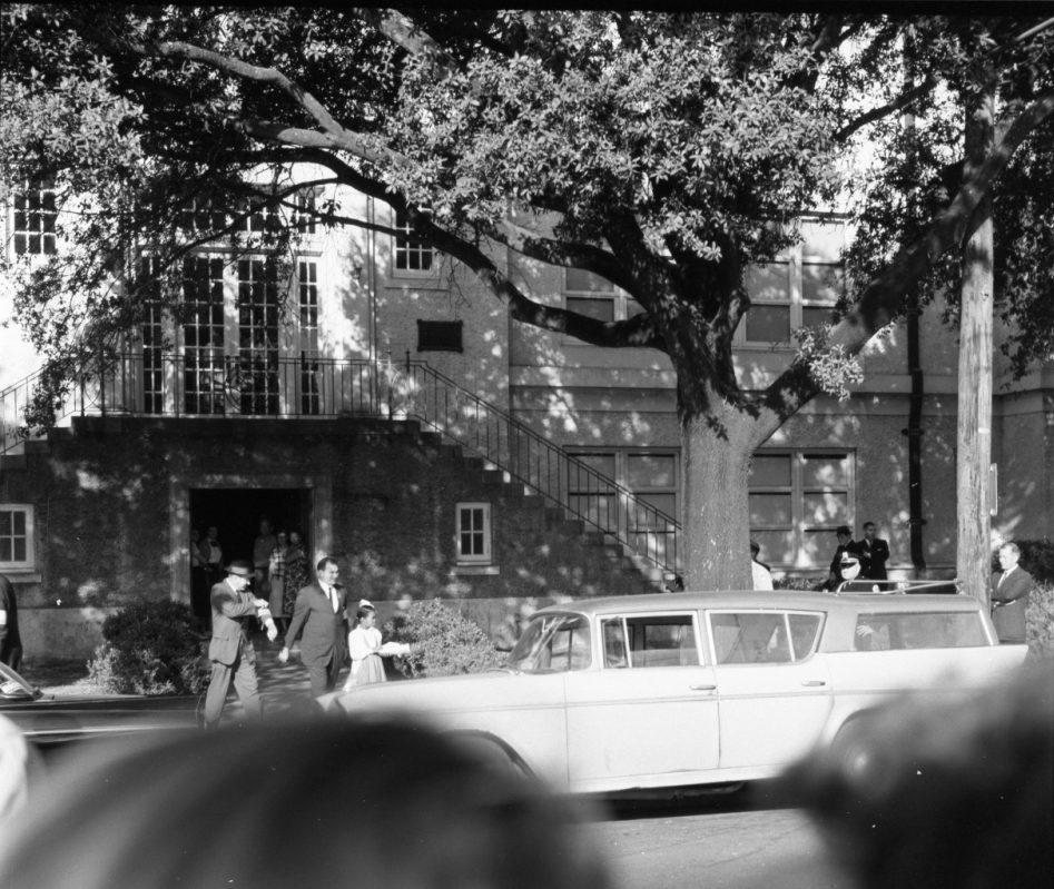 A vintage photograph shows a scene outside a building with a set of stairs and large trees. People are gathered near a parked classic station wagon, and more individuals are seen near the entrance and windows of the building.