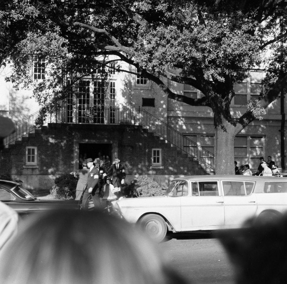 A black and white photo of a vintage car parked on a street in front of a building. People are gathered around, some on the steps leading to the entrance, with a large tree casting shadows on the scene.