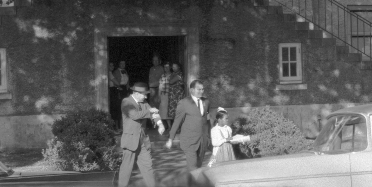 A vintage photo shows a man in a suit and hat leading another man in a suit and a young girl holding a book out of a building. More people stand in the doorway. A car is parked in front. The scene is in black and white.