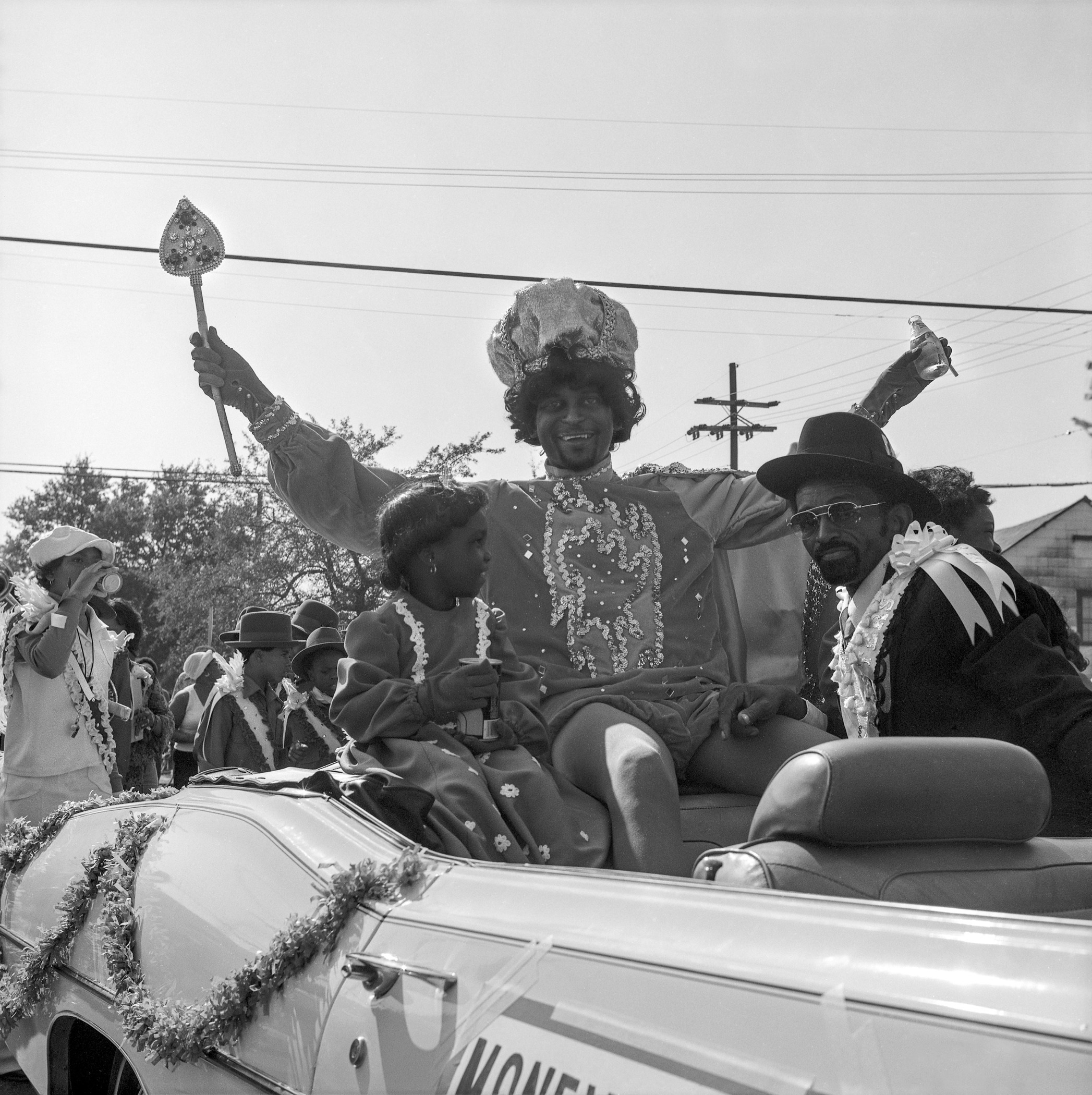 People dressed in festive costumes ride on the back of a decorated convertible during a parade. A man in an elaborate outfit holds a scepter, surrounded by others in hats and leis, with power lines and trees in the background.