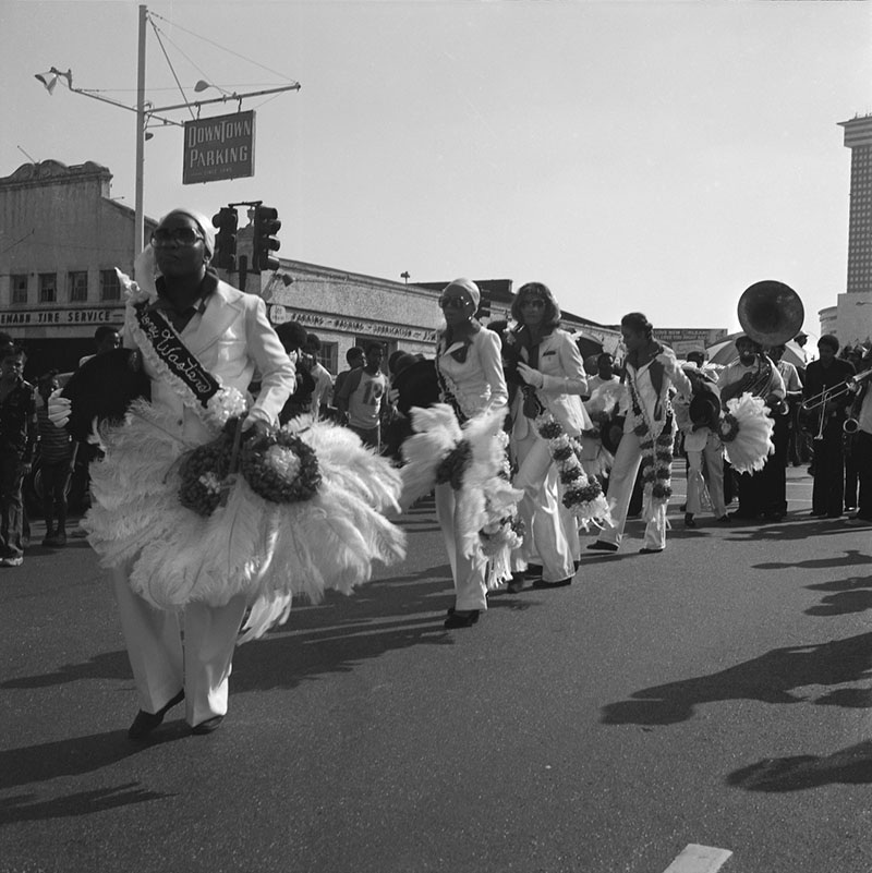 A black and white photo of a parade featuring a group of people in formal attire adorned with fluffy feathered accessories. They are marching with musical instruments on an urban street, with a Downtown Parking sign visible in the background.